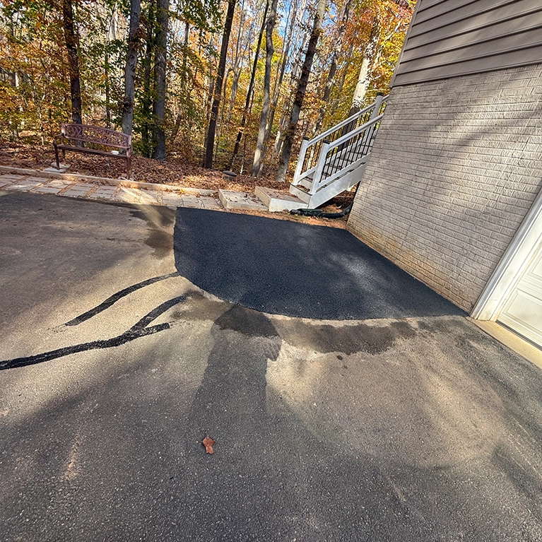 Freshly paved asphalt area next to a garage, with a stairway in the background and a wooden bench amid autumn foliage.